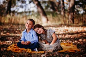 Two children sitting on orange blanket in fallen autumn leaves during outdoor photo session.