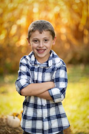 Child in blue plaid shirt smiles confidently with arms crossed against autumn background.