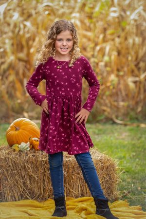 Young person in burgundy dress poses by hay bale and pumpkin in fall setting.