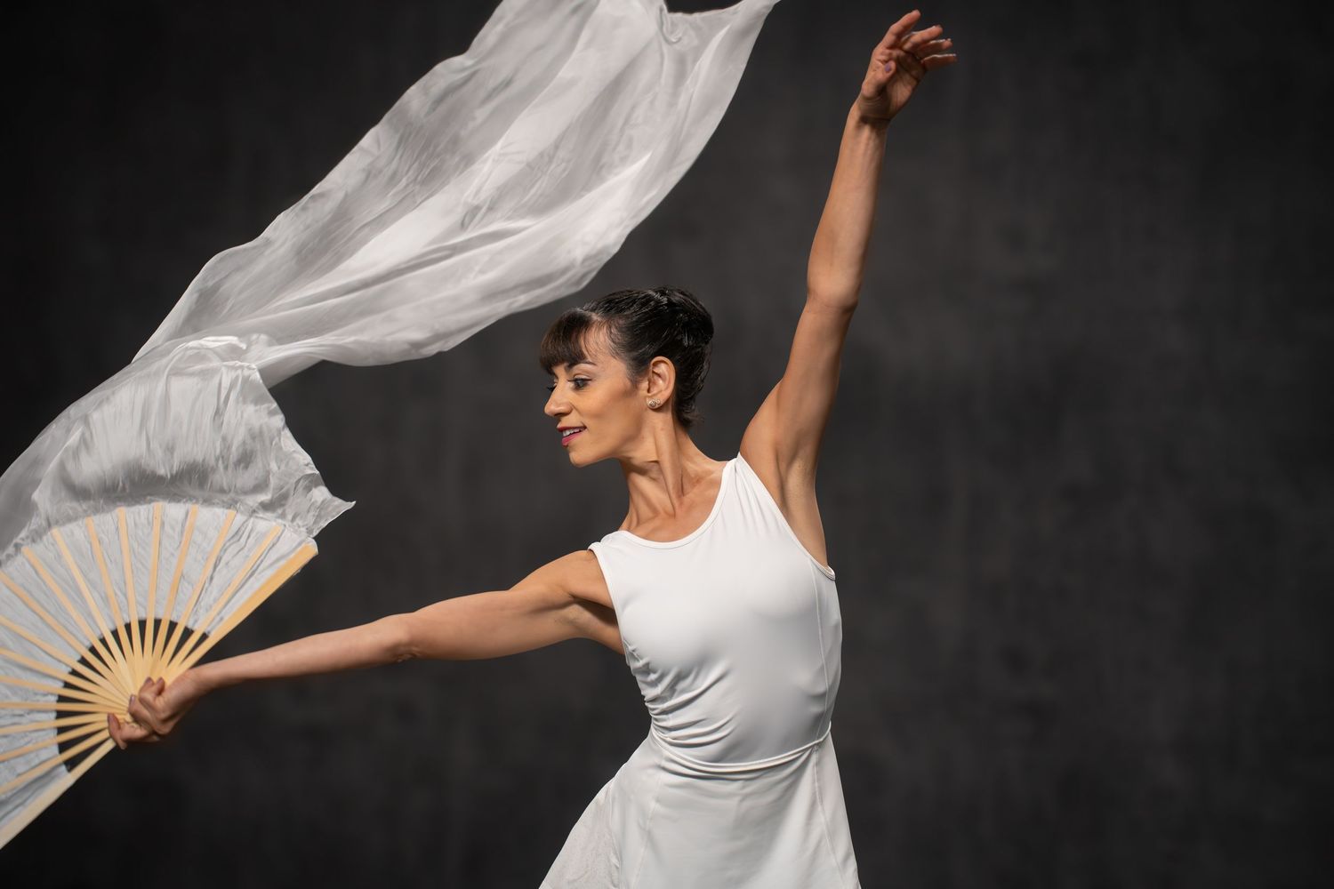 Ballet dancer in white costume performs graceful movements with flowing fabric against dark background.