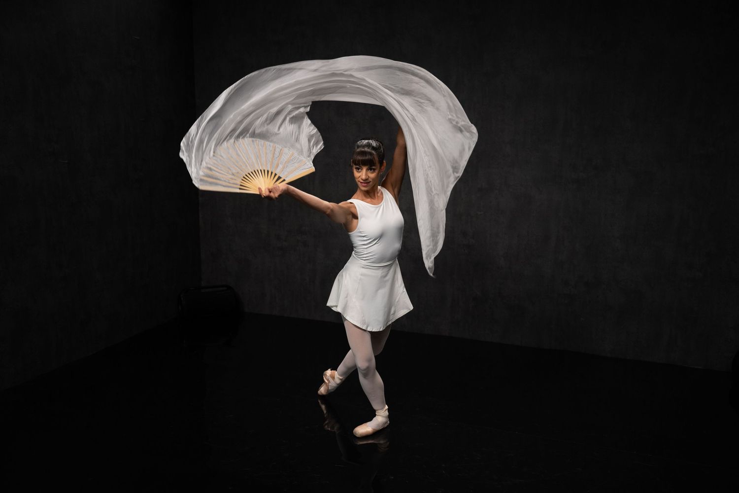 Dancer in white leotard creates elegant shapes with flowing fabric in dramatic studio lighting.