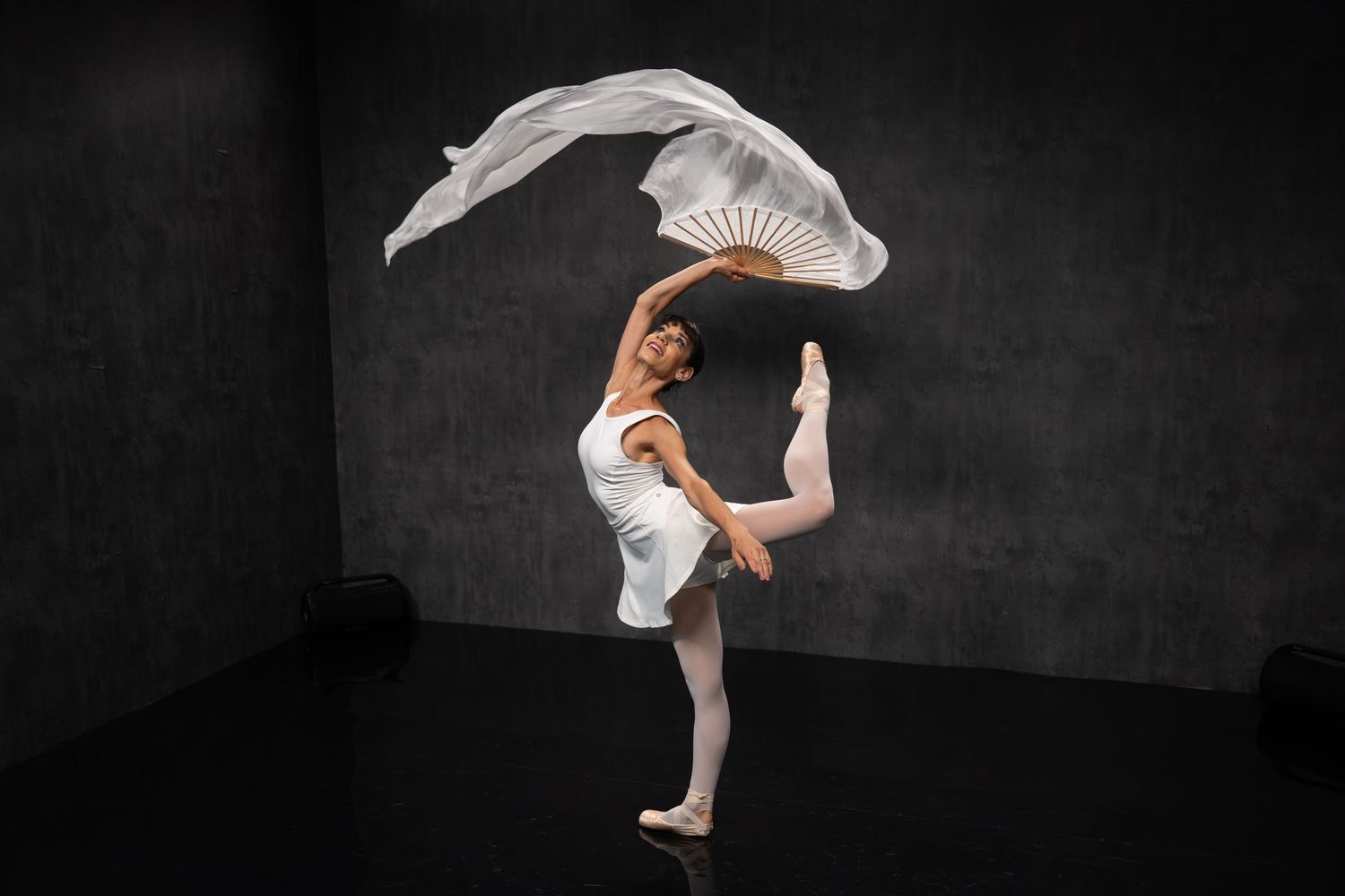 Contemporary dancer moves expressively with billowing white fabric in moody black studio setting.