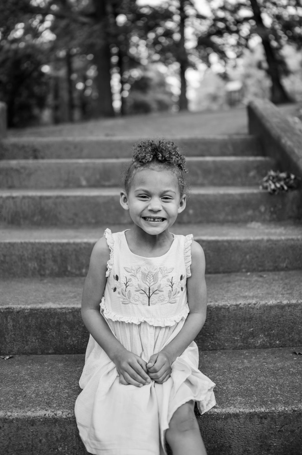 A black and white portrait of a child sitting on concrete steps wearing a white dress.