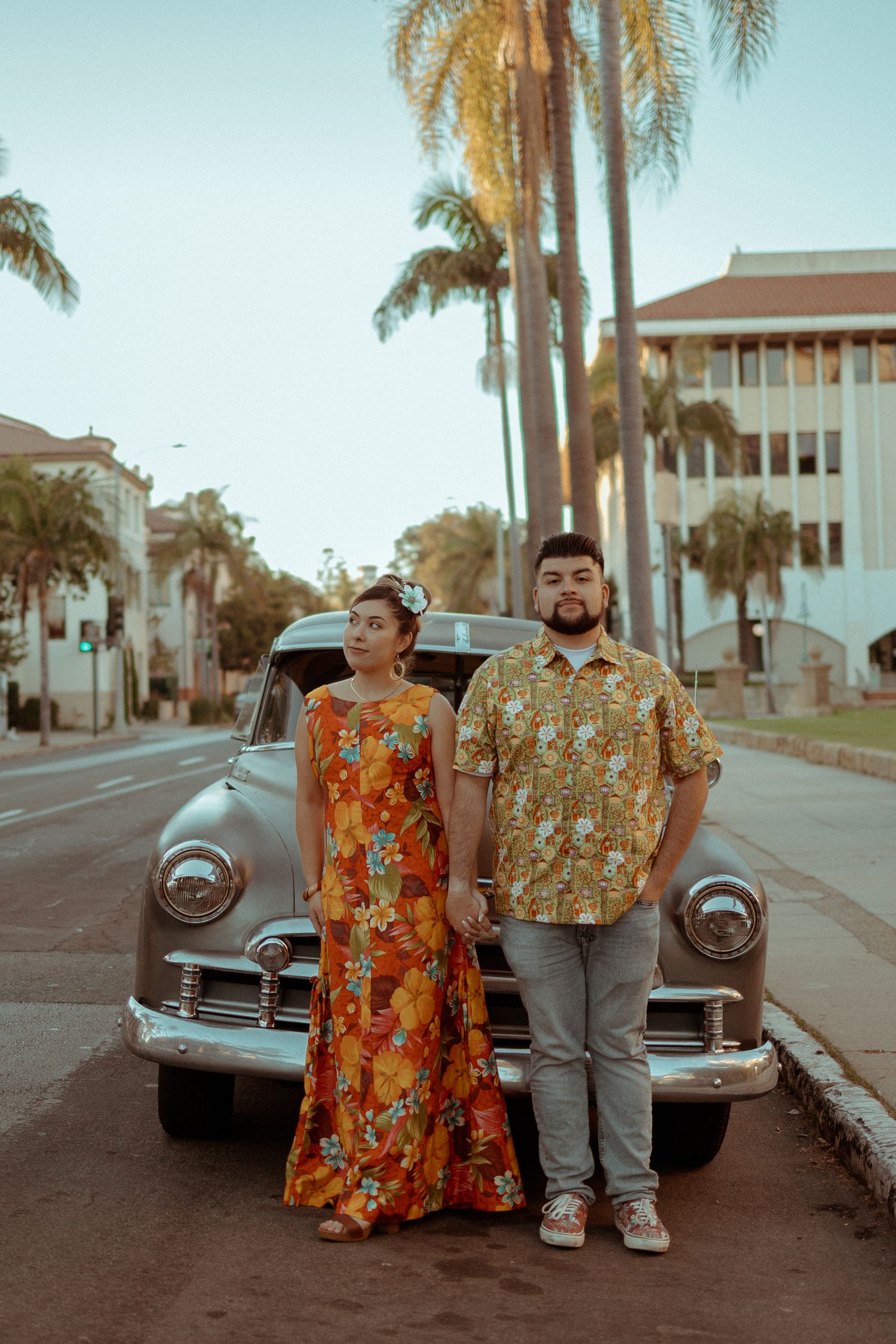 Vintage car parked on palm tree-lined street with couple in colorful floral outfits posing beside it at sunset.