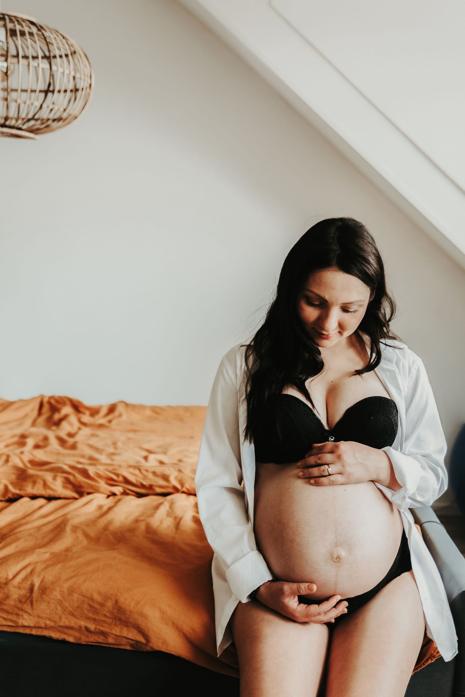 Intimate maternity photo of an expectant mother in black lingerie and white shirt against orange bedding.