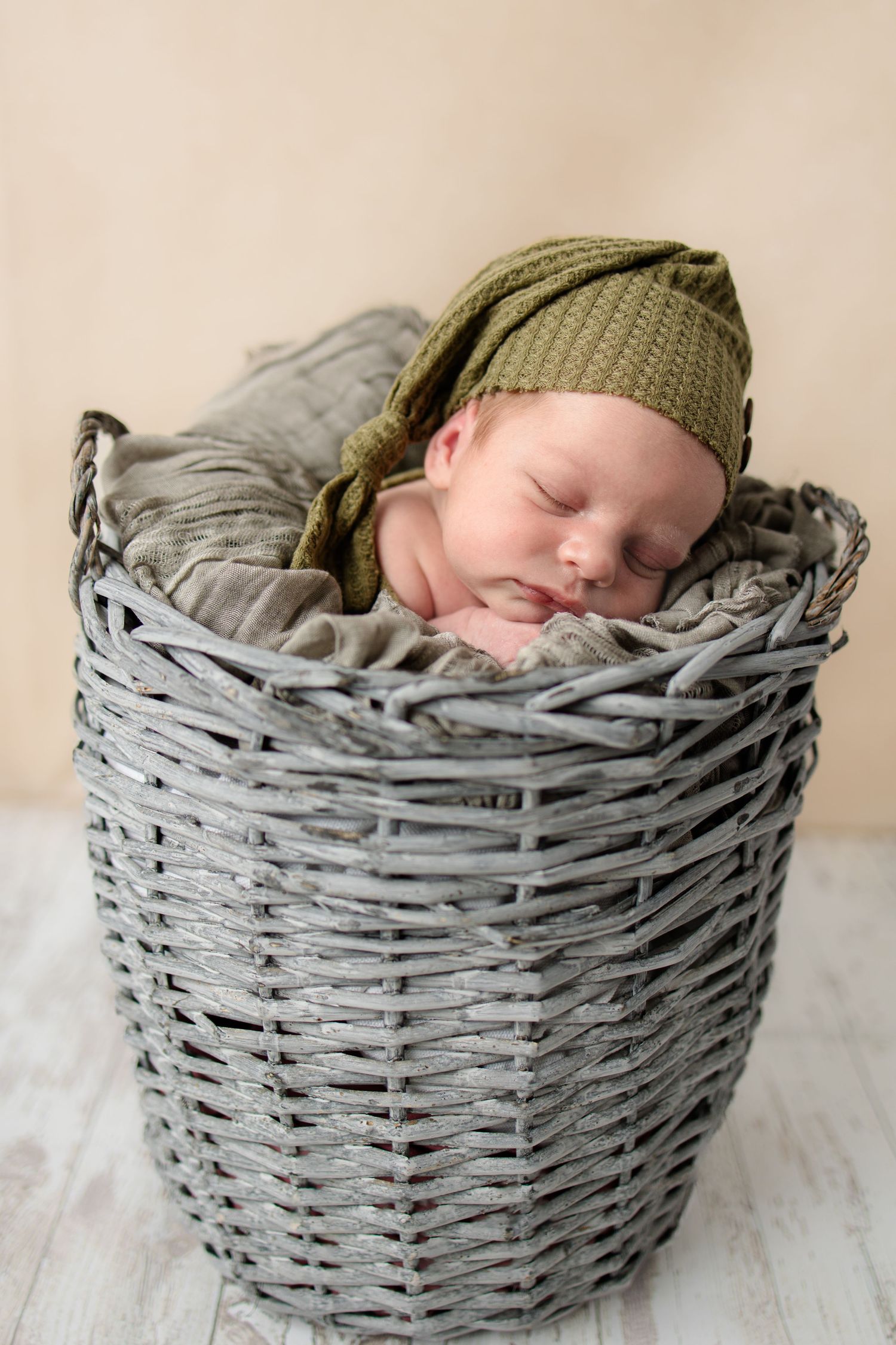 Sleeping newborn wearing green knit hat nestled in gray woven basket.