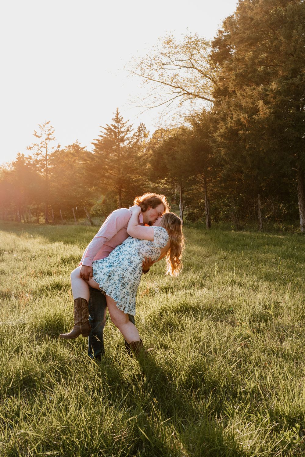 A romantic dip pose in a sunlit field with pine trees in the background.