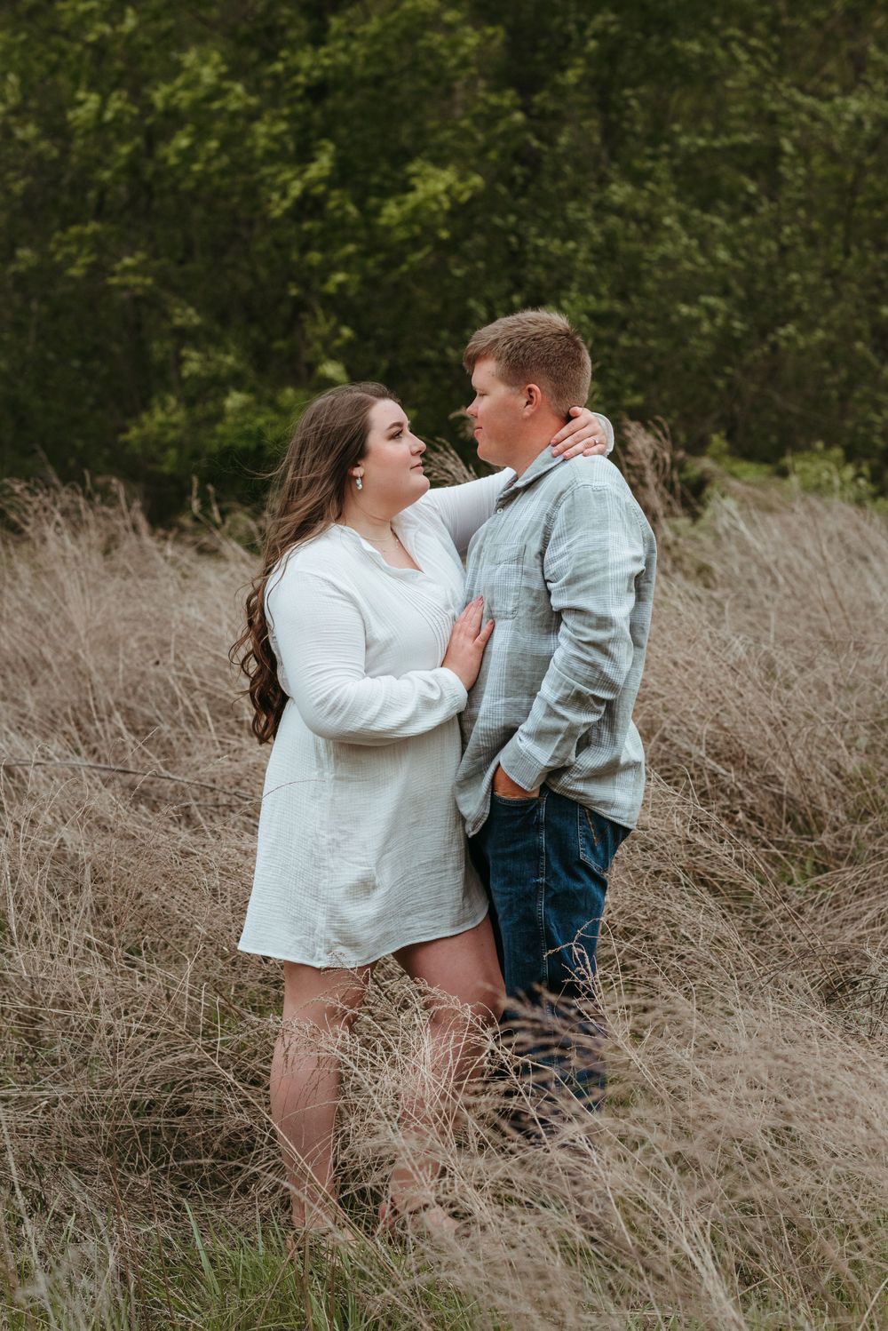 A couple embraces in a field of dried grass with trees in the background.