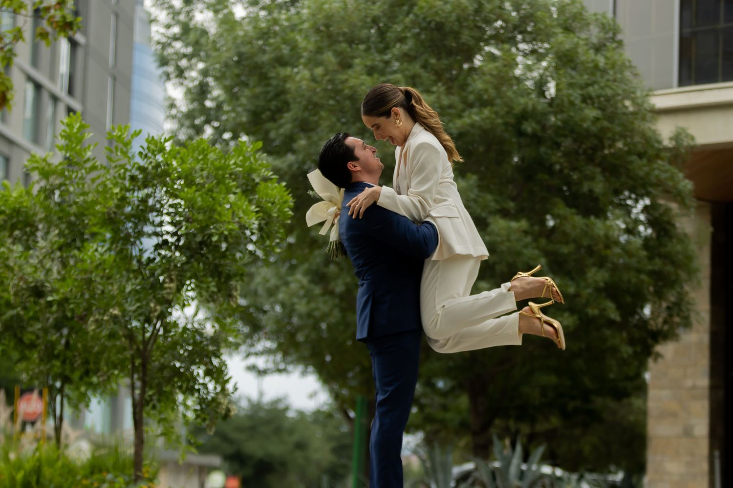 Man lifting woman in white suit joyfully outdoors among green trees.