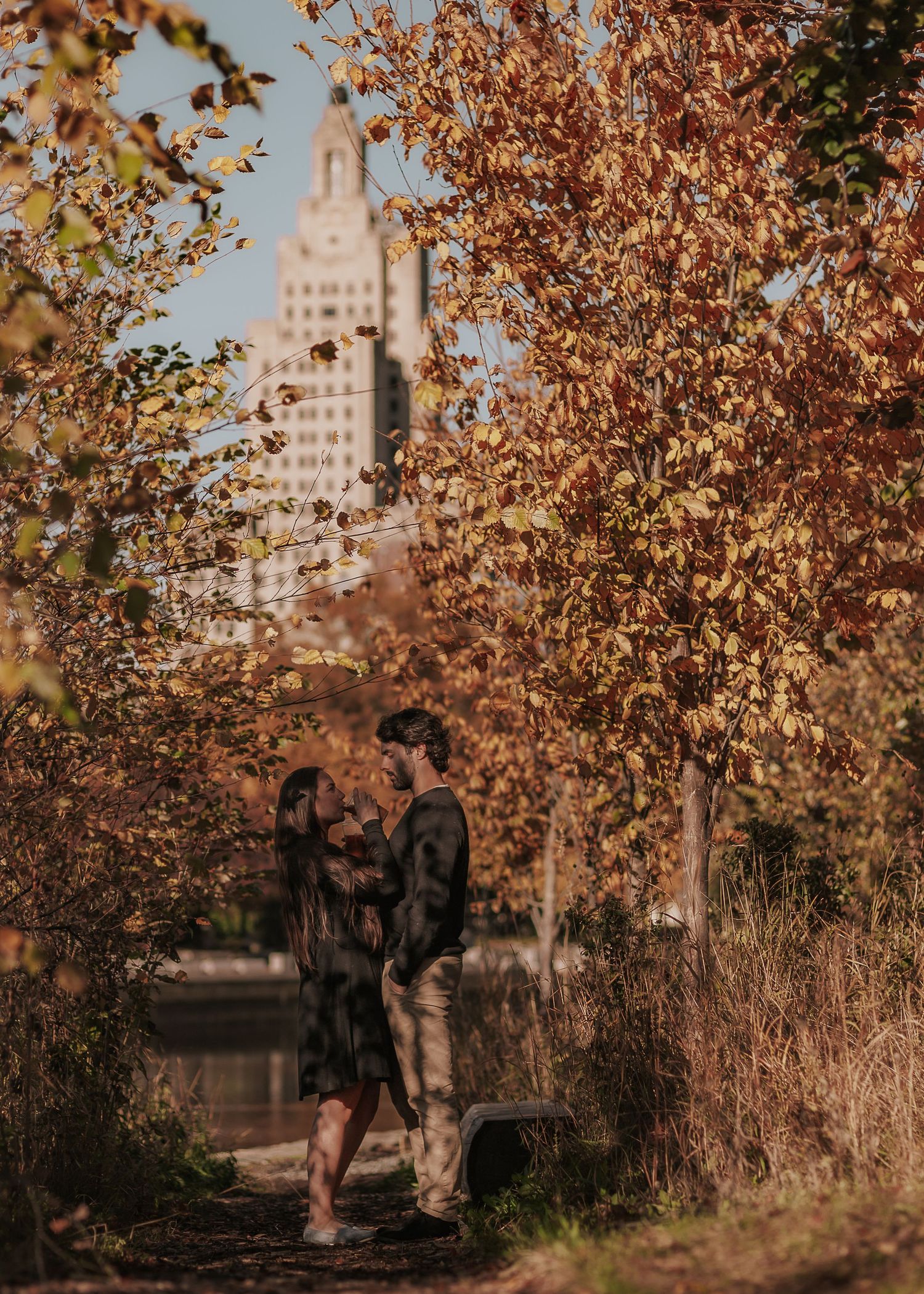Autumn trees with golden leaves frame a scenic urban pathway along a riverfront with city buildings in the background.