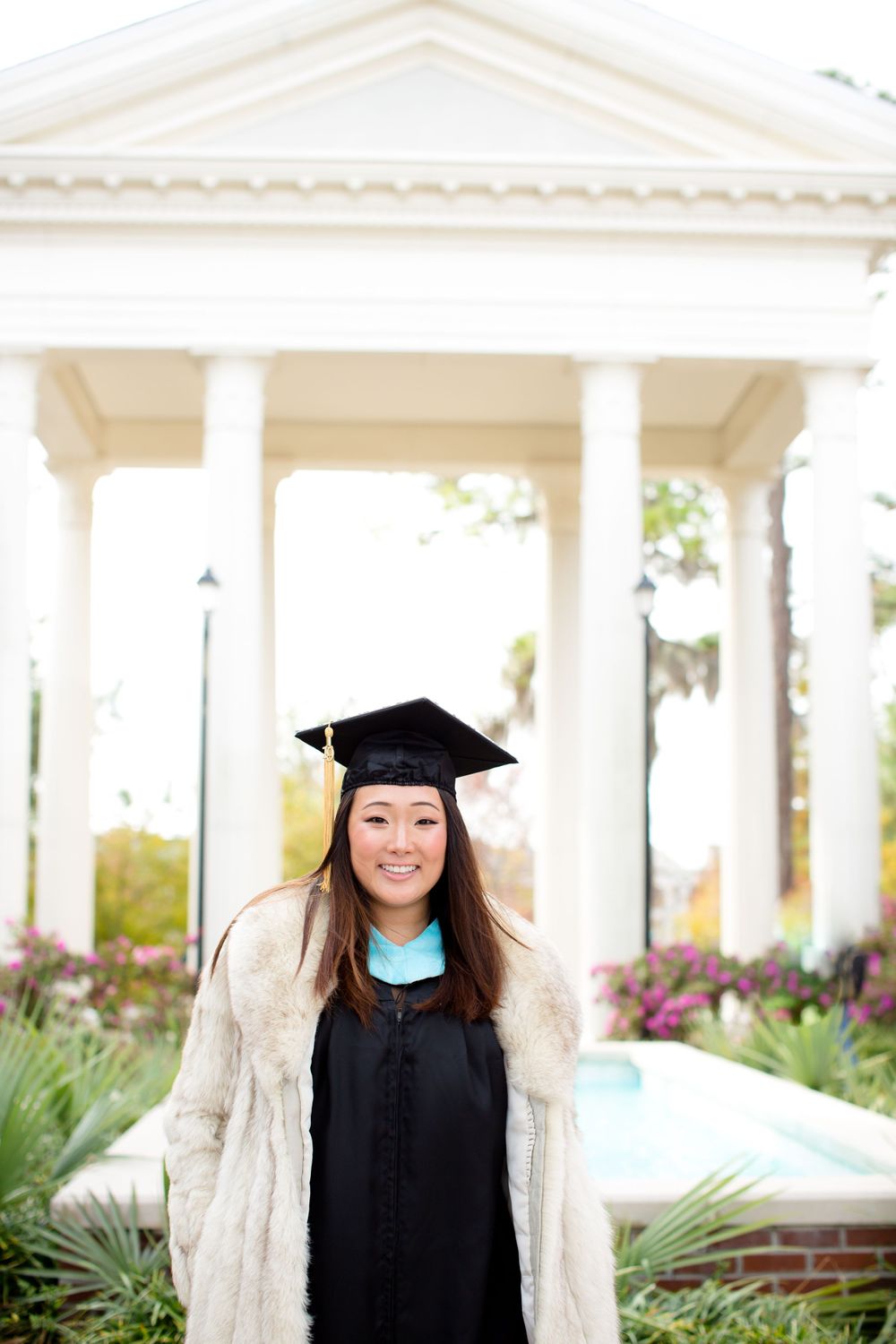Graduate in black gown and fur coat smiling in front of classic white gazebo.