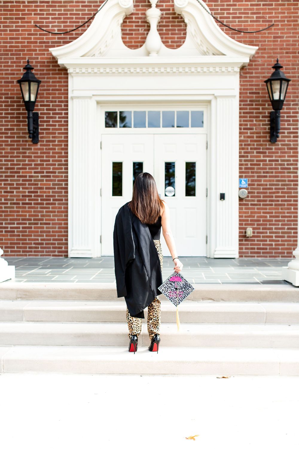 Figure in leopard print pants and black coat stands before elegant white doorway with ornate trim and lanterns.