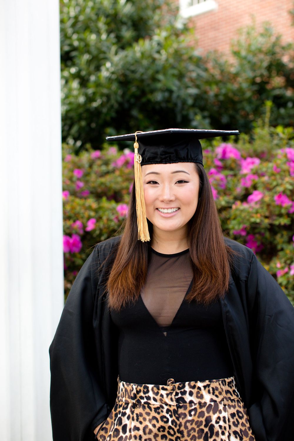 Graduate wearing cap and gown with leopard print skirt posing by flowering bushes.