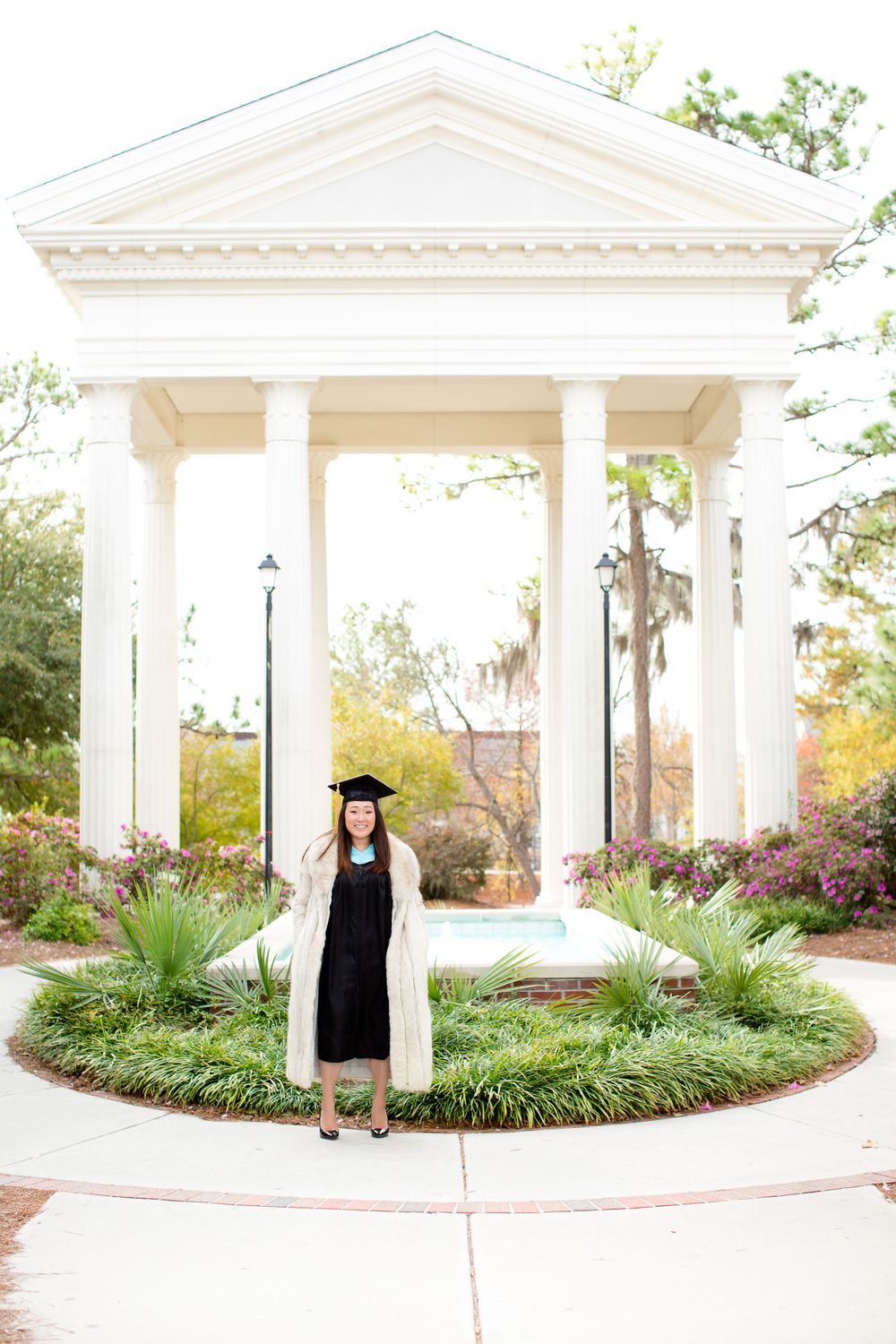 Graduate in cap and gown standing in front of white columned gazebo with landscaped garden.