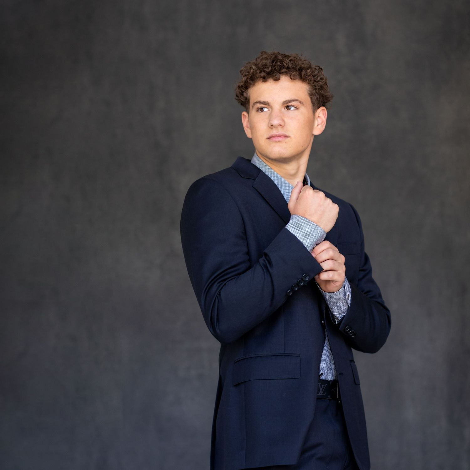 Professional studio portrait of a high school senior in a navy blue suit adjusting their cuff against a dark gray background.