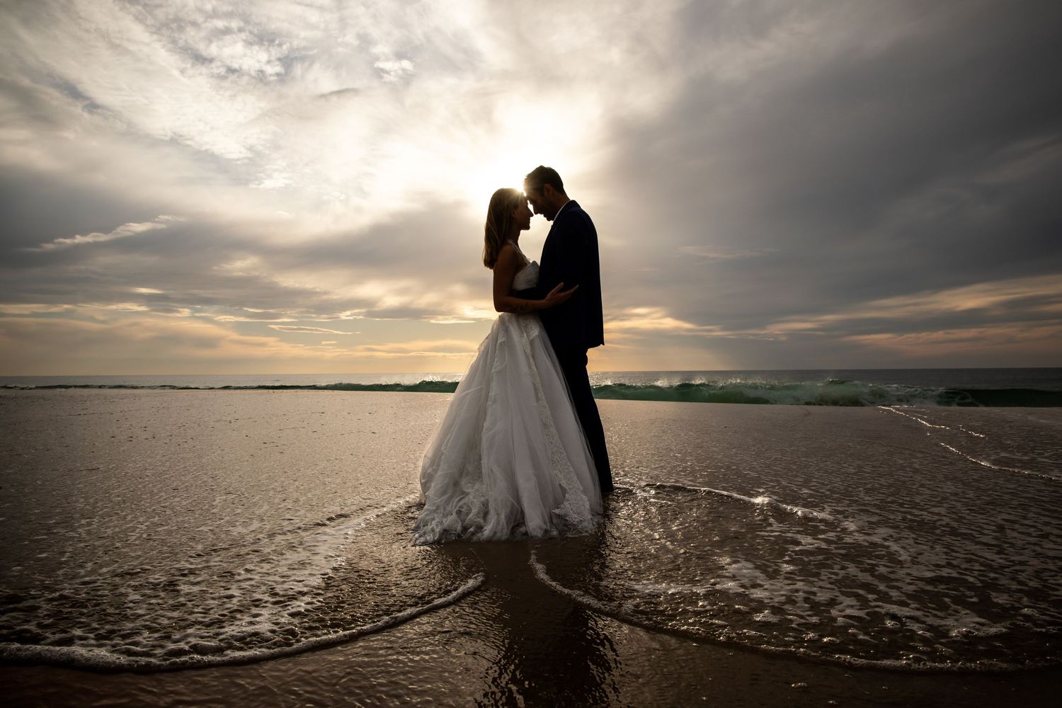 Romantic bride and groom silhouette kissing on beach at sunset with dramatic sky.