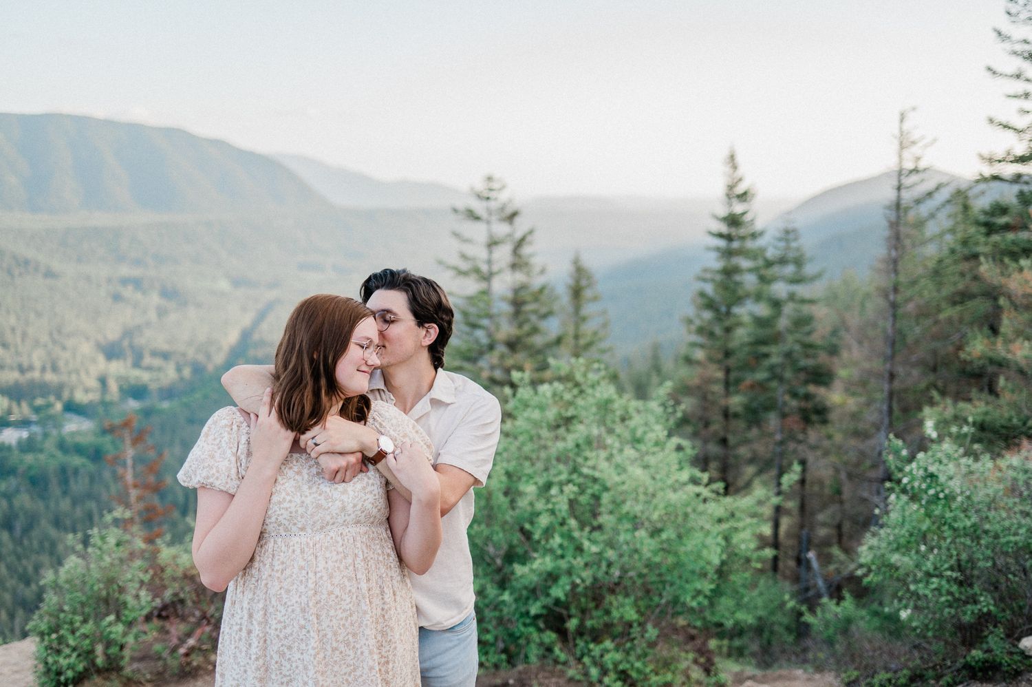 A romantic couple shares a tender moment in a mountain forest setting with evergreen trees and misty hills in the background.