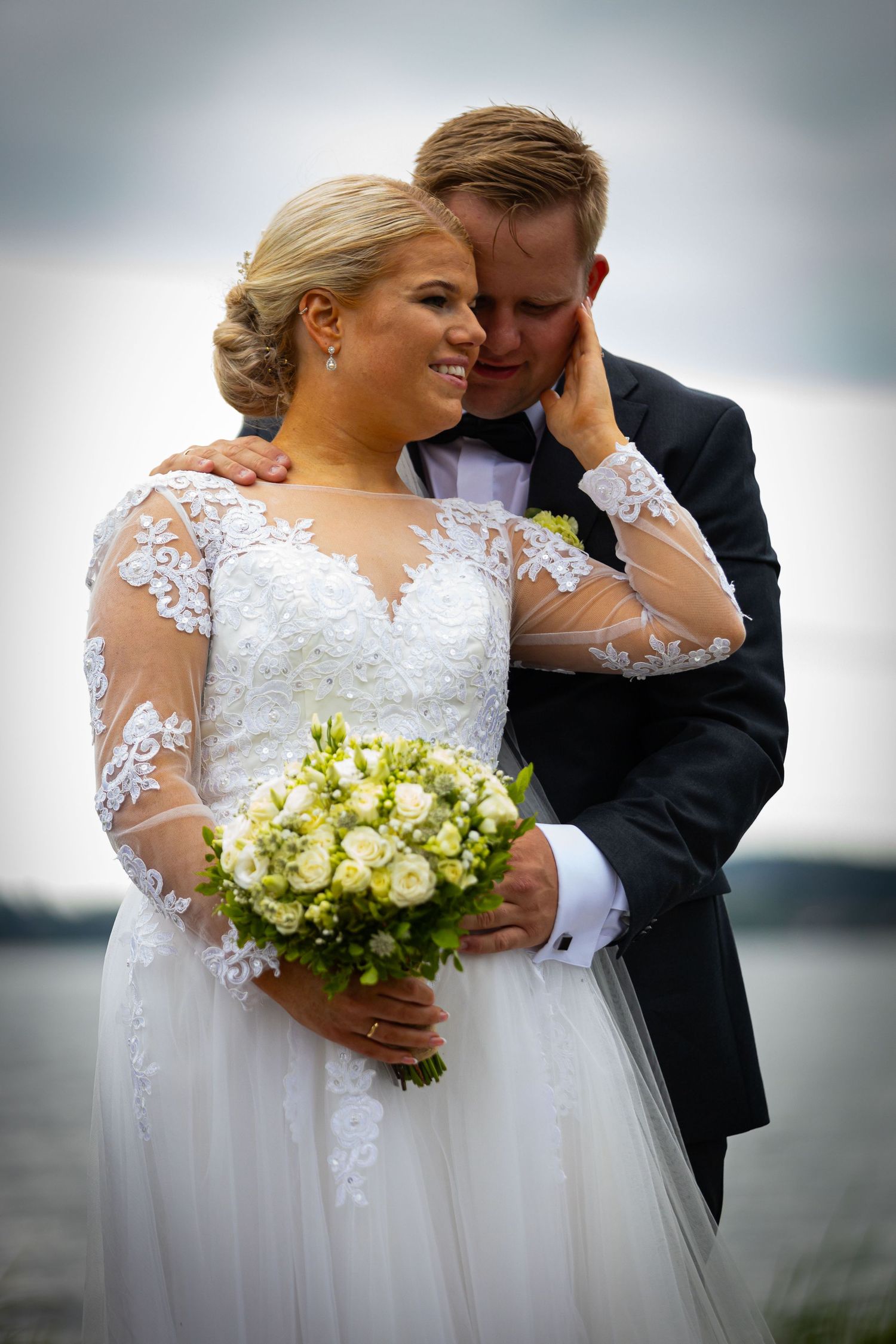 Smiling bride and groom embracing outdoors by a lake, bride holding white and green bouquet.