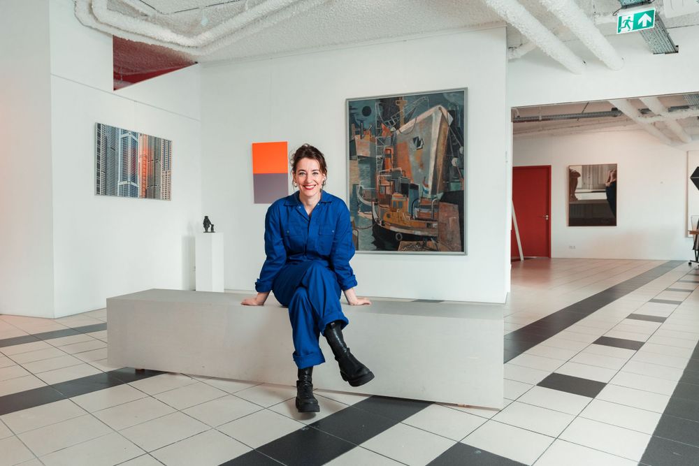 A figure in bright blue attire sits in a modern art gallery with white walls and geometric flooring.