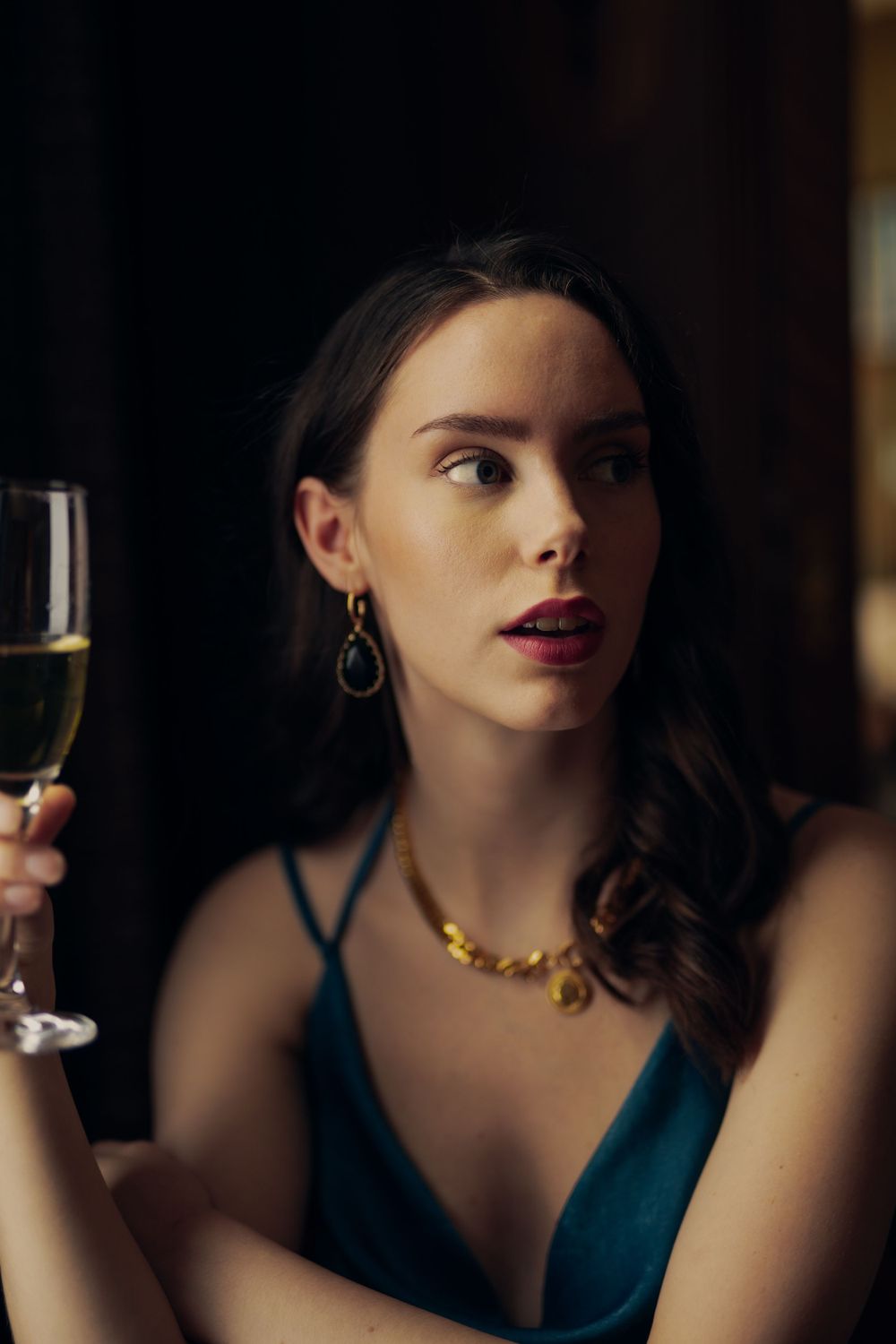 Elegant figure in teal dress holds champagne glass in dramatic evening lighting.