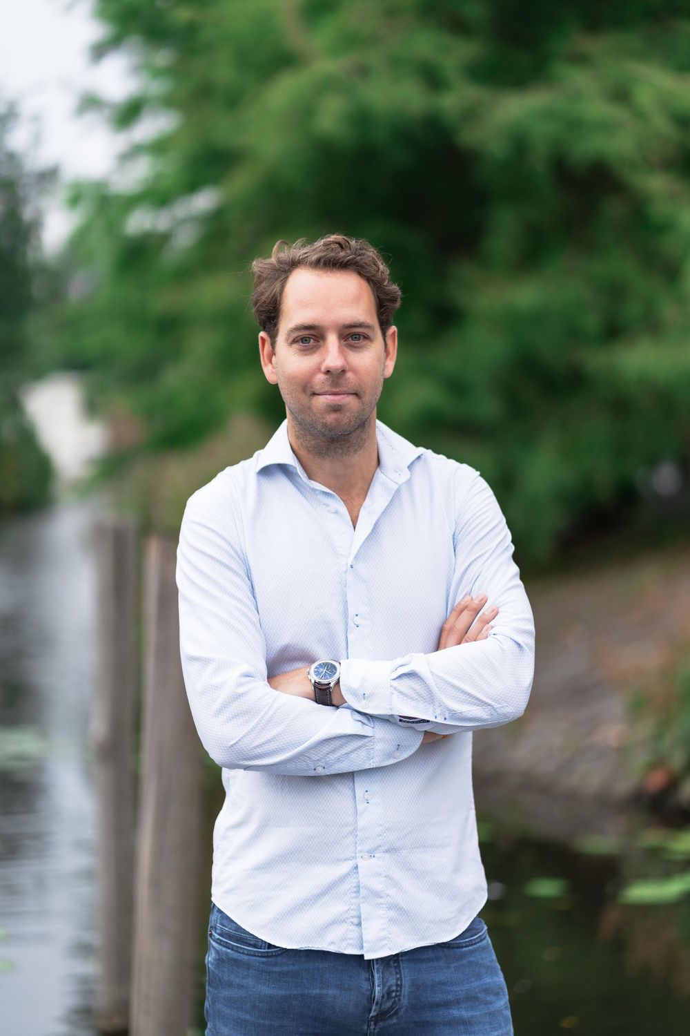 Professional wearing a white button-down shirt stands with crossed arms outdoors near greenery.