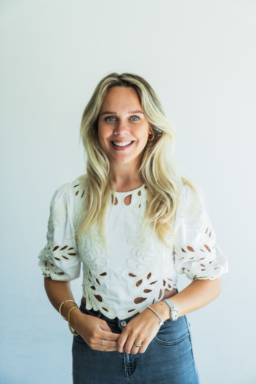 Individual in white eyelet top and jeans poses against white background with natural lighting.