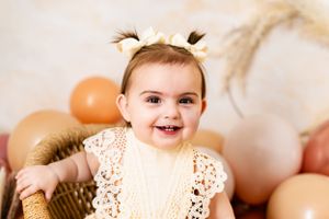 Baby with lace dress lying among peach and neutral colored balloons.