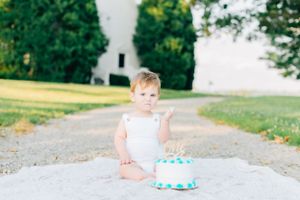 A baby in white sits on a pathway with a small birthday cake.