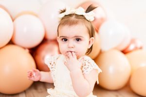 Smiling baby in white lace outfit posing with balloon decorations.