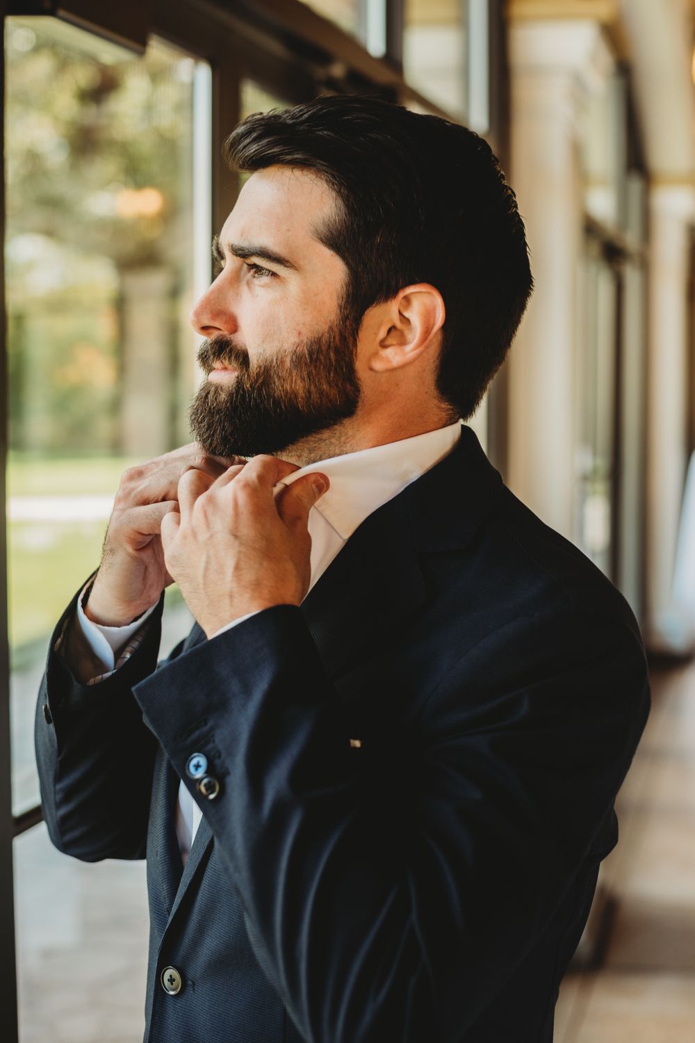 Profile view of person in dark suit adjusting collar near window.