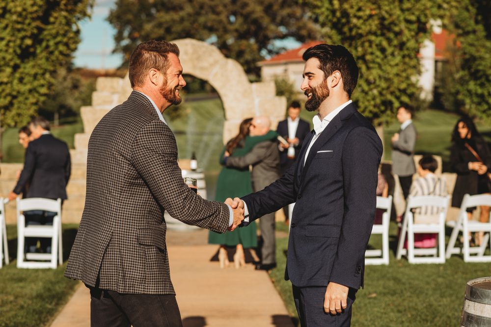 Two people in suits greet each other warmly at an outdoor wedding ceremony venue.