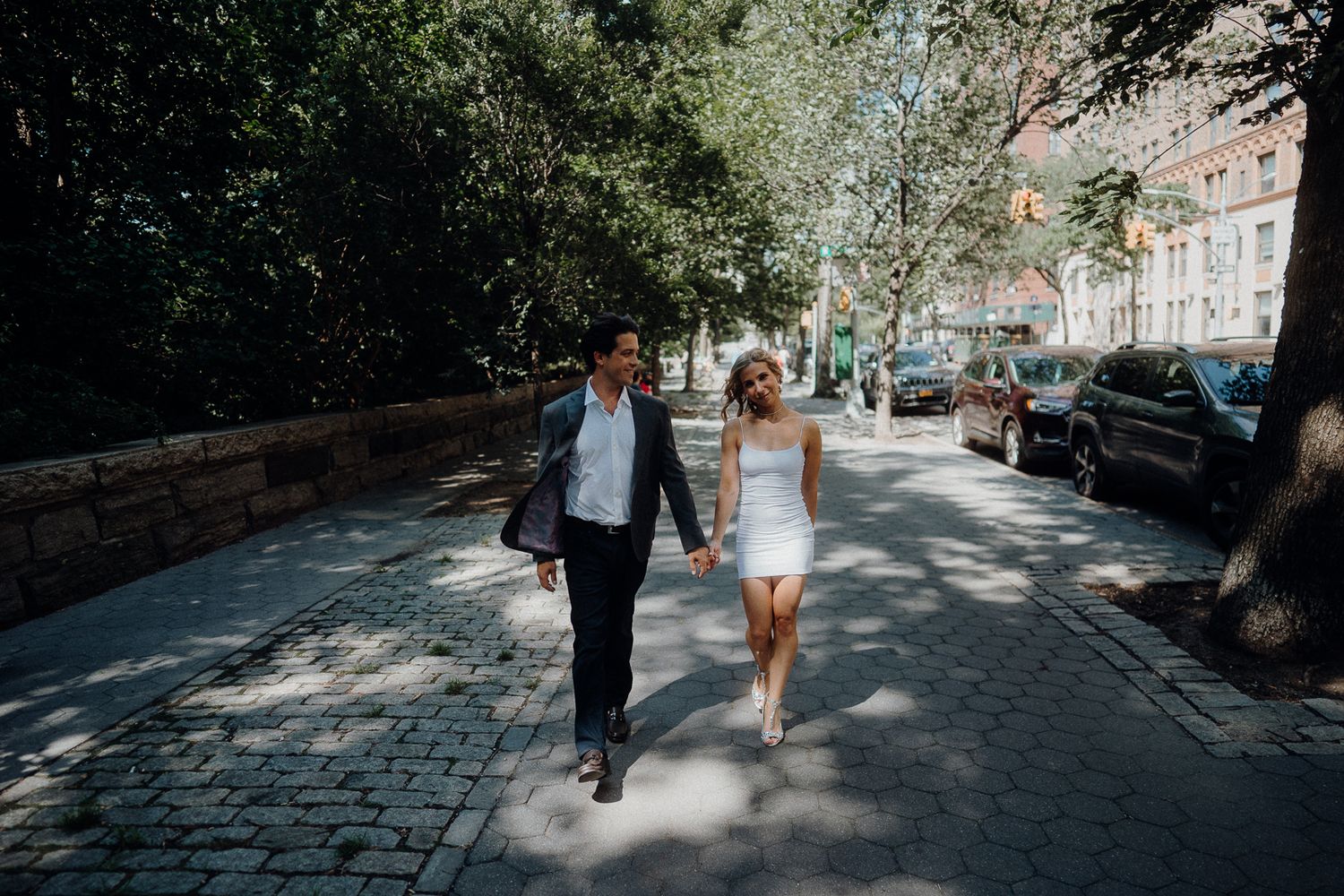 A couple walks together on a tree-lined cobblestone street in an urban setting.