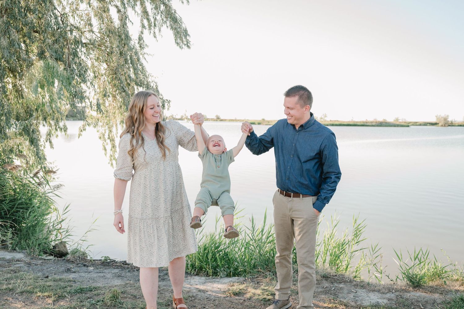 Parents swing child between them by lakeside under willow branches.