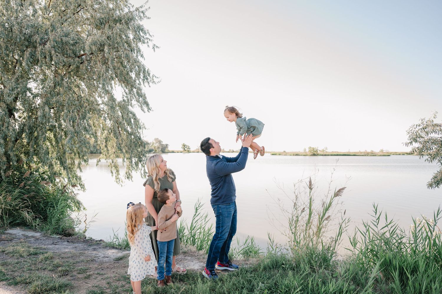 Family plays together by lake shore during outdoor photo session.