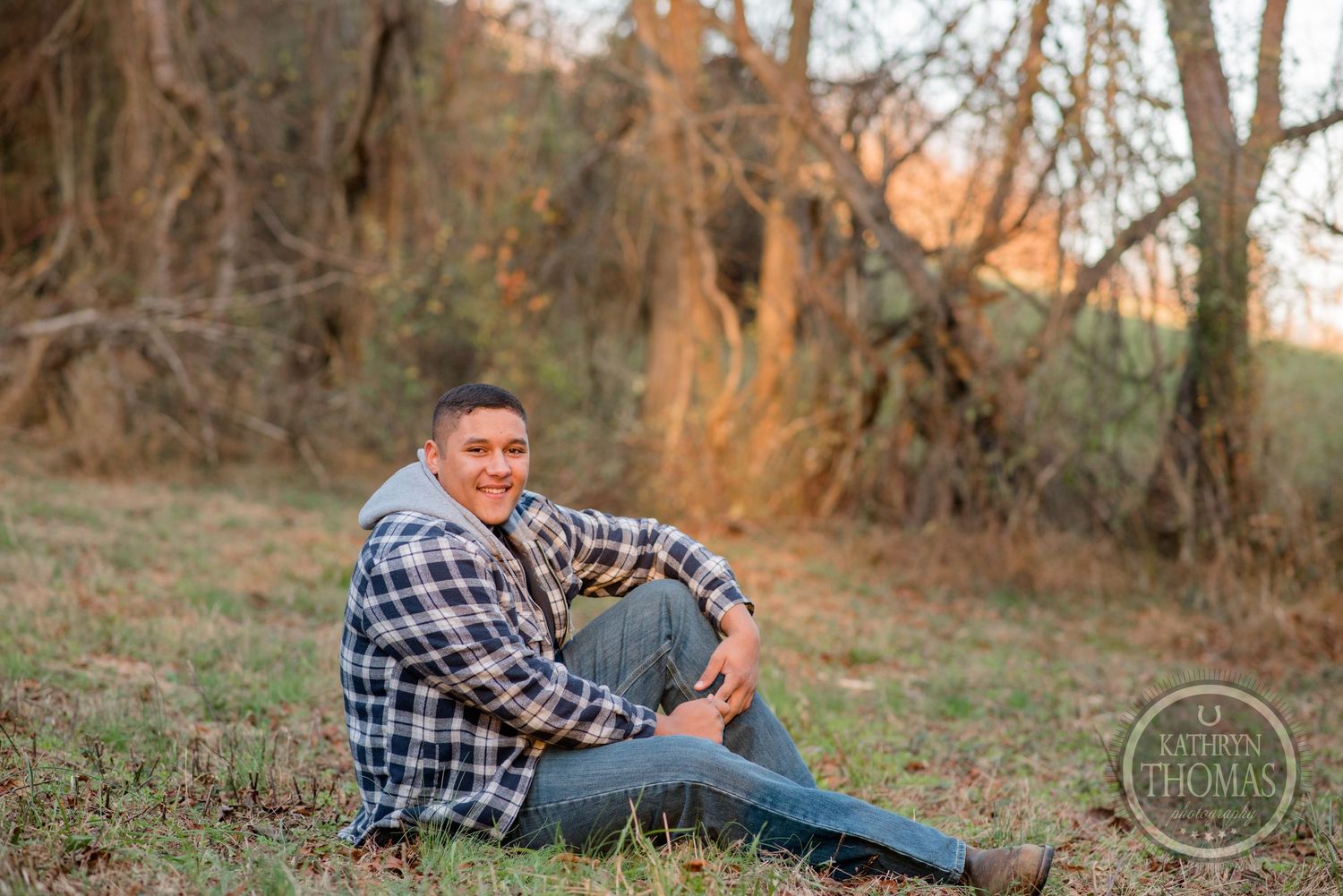 A person in plaid and jeans sits casually on grass with fall foliage in the background.