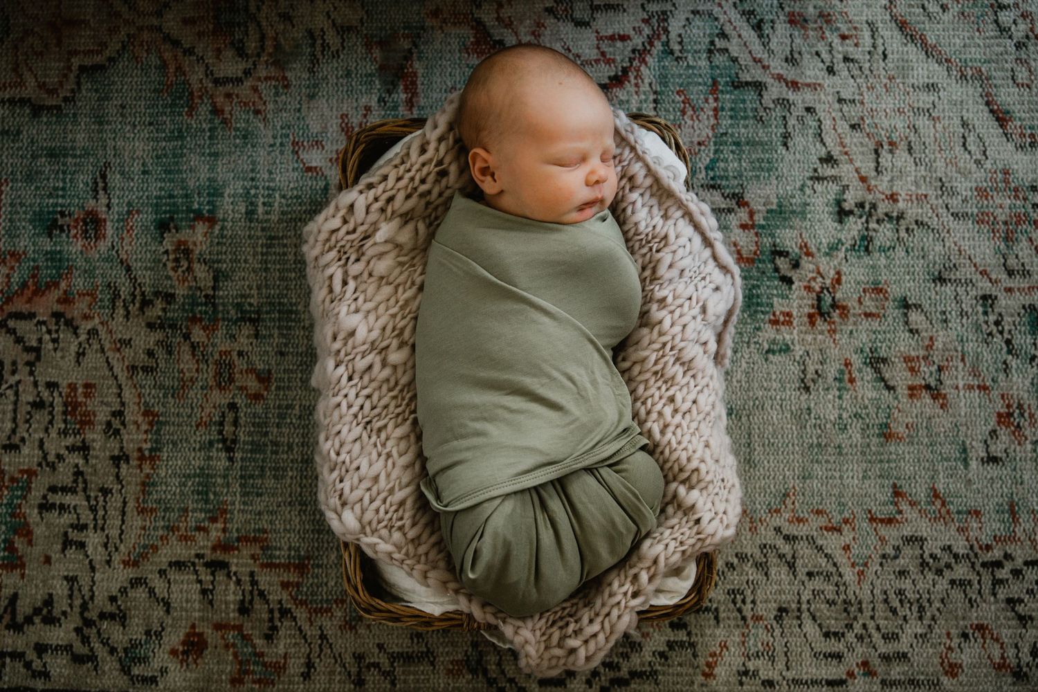Sleeping posed newborn baby swaddled in sage green wrap, nestled in a basket with chunky knit blanket.