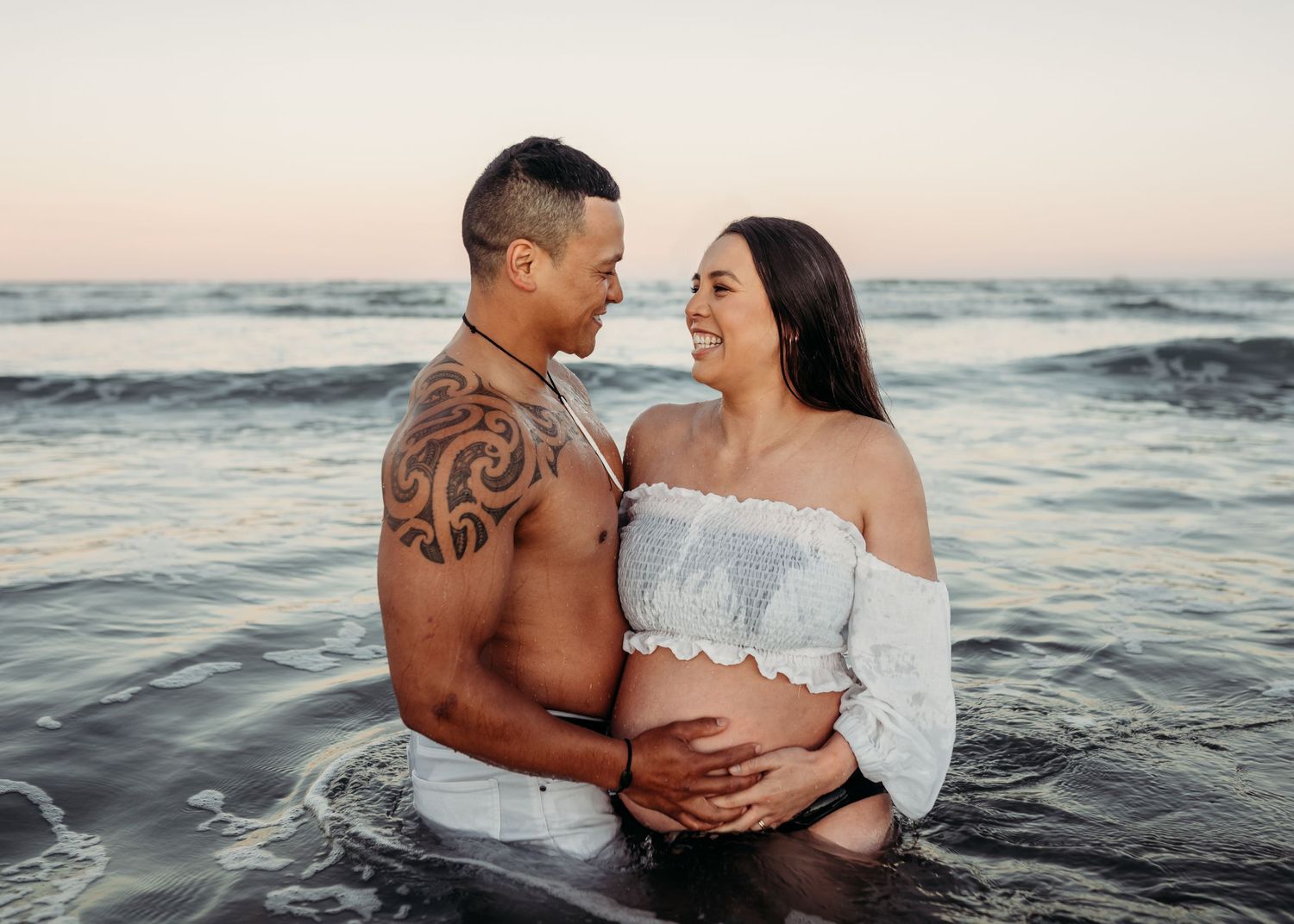 Intimate maternity photo of couple embracing in ocean waves at sunset.