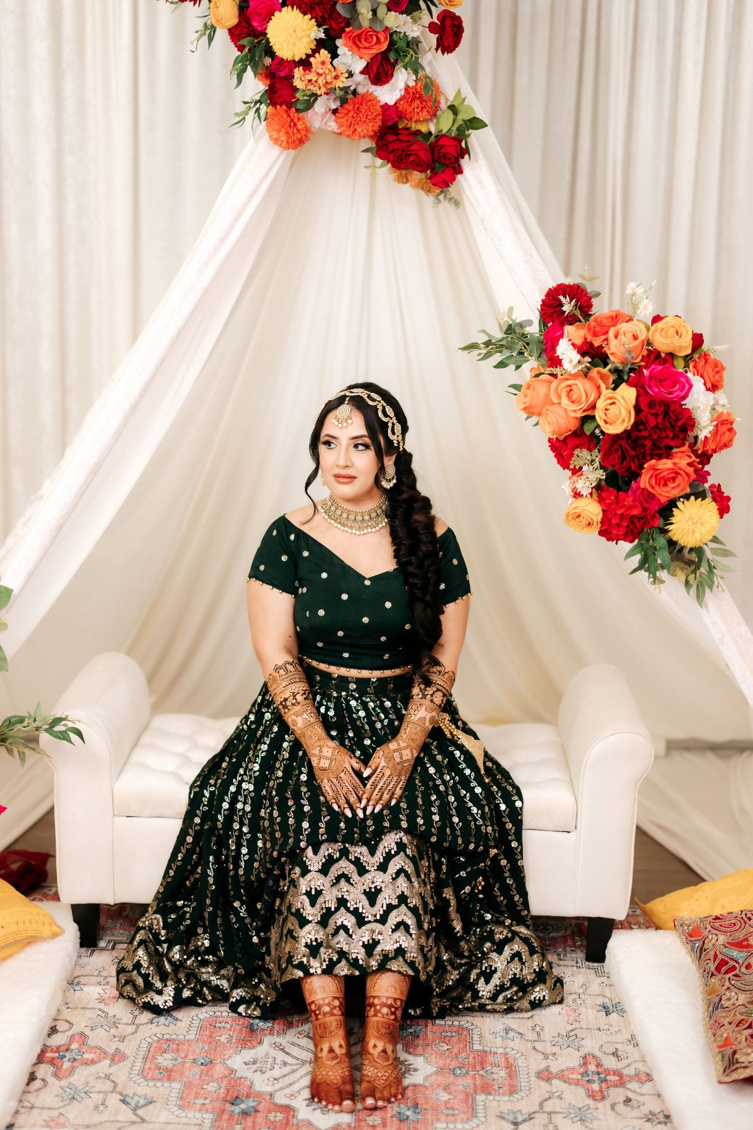 Traditional South Asian bride in emerald green lehenga seated under floral arch decor.