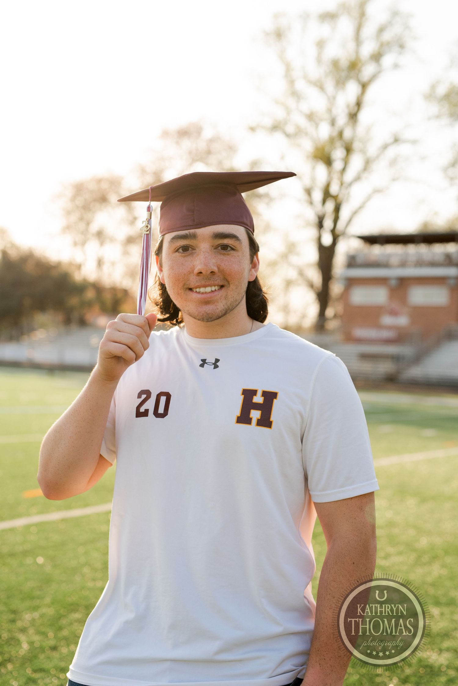 Graduate in cap and white jersey holds tassel while posing for outdoor senior photo on sports field.