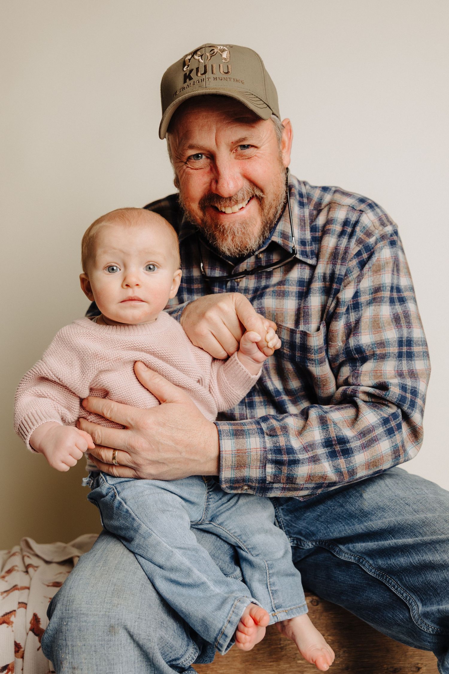 A tender family portrait showing an adult in a plaid shirt and baseball cap holding a baby in a pink sweater.