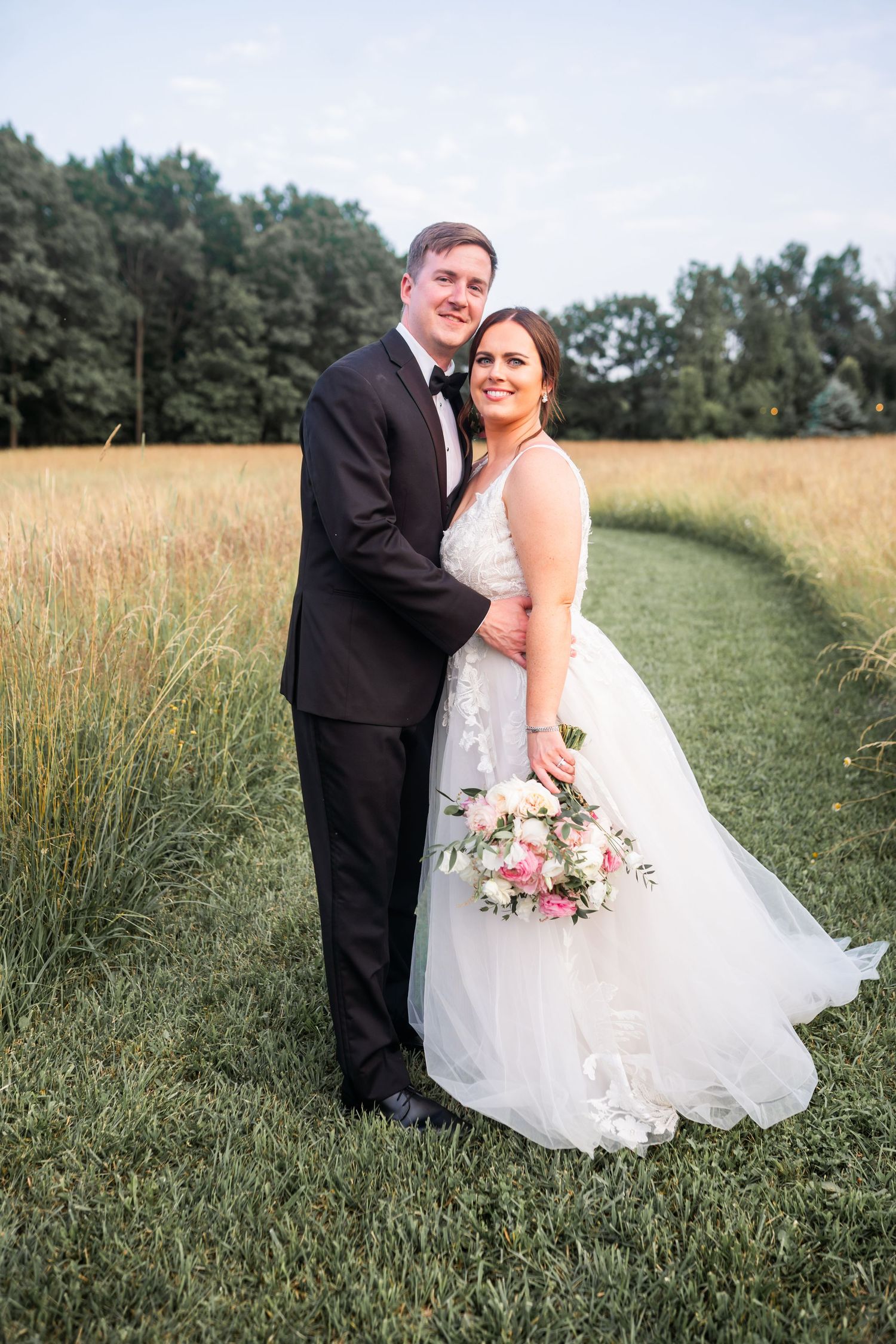 A bride and groom pose together in a grassy field, the bride wearing a white wedding dress and the groom in a black suit.