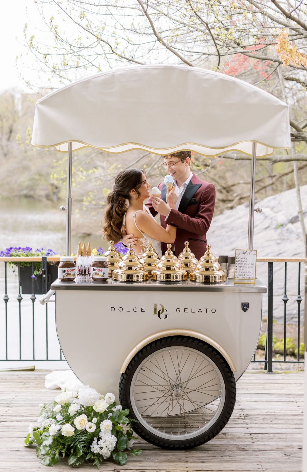 Couple enjoys gelato from vintage ice cream cart decorated with flowers at outdoor wedding venue.