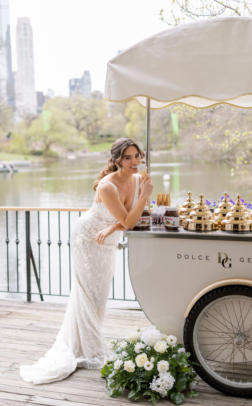 Bride in lace gown enjoys gelato beside vintage cart with Central Park skyline in background.