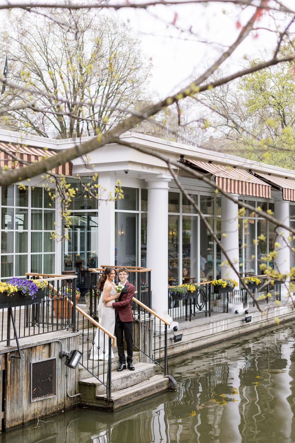 A couple stands on a waterfront terrace with white columns and striped awnings overlooking a pond.