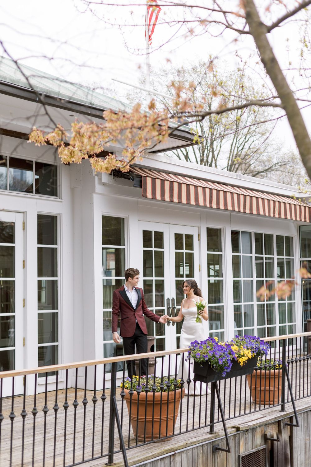 A couple on an elegant waterfront terrace with striped awnings and purple flowers in planters.