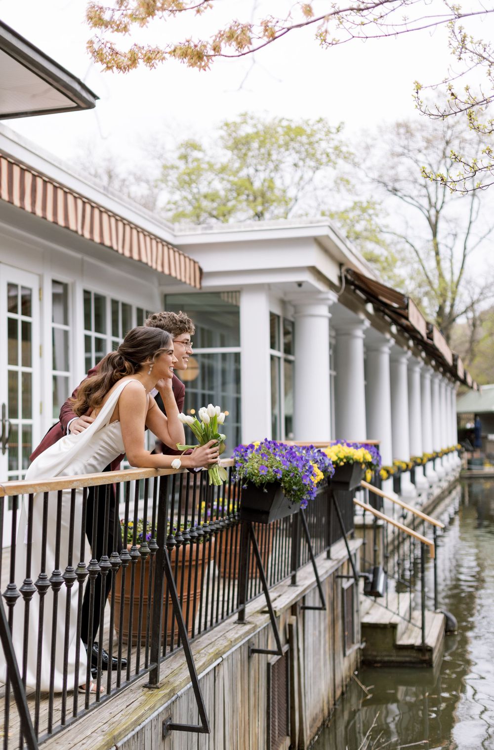 Two people lean on a railing at a waterfront venue with flower boxes and white columns.