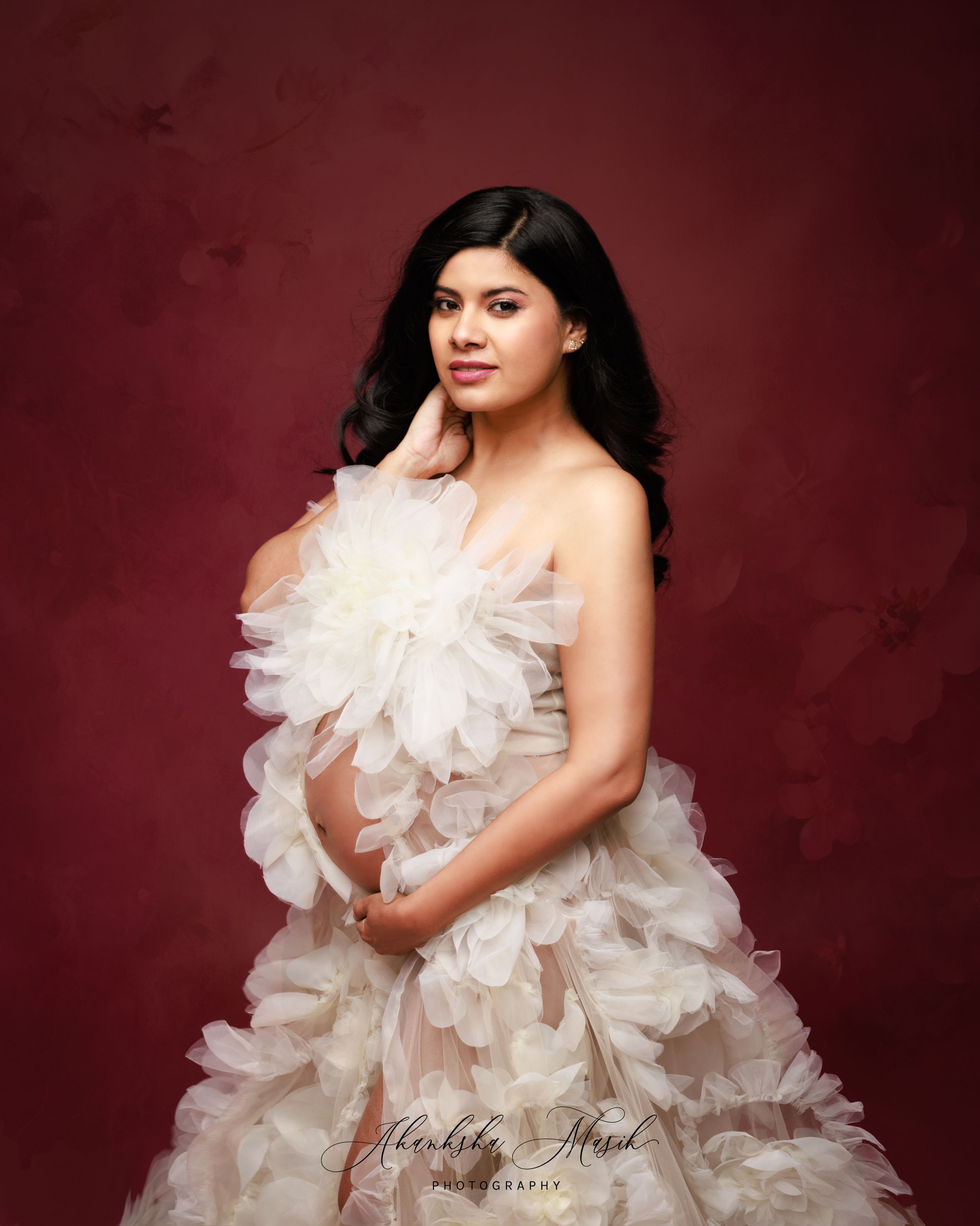 Elegant maternity studio portrait with voluminous white tulle dress against burgundy backdrop.