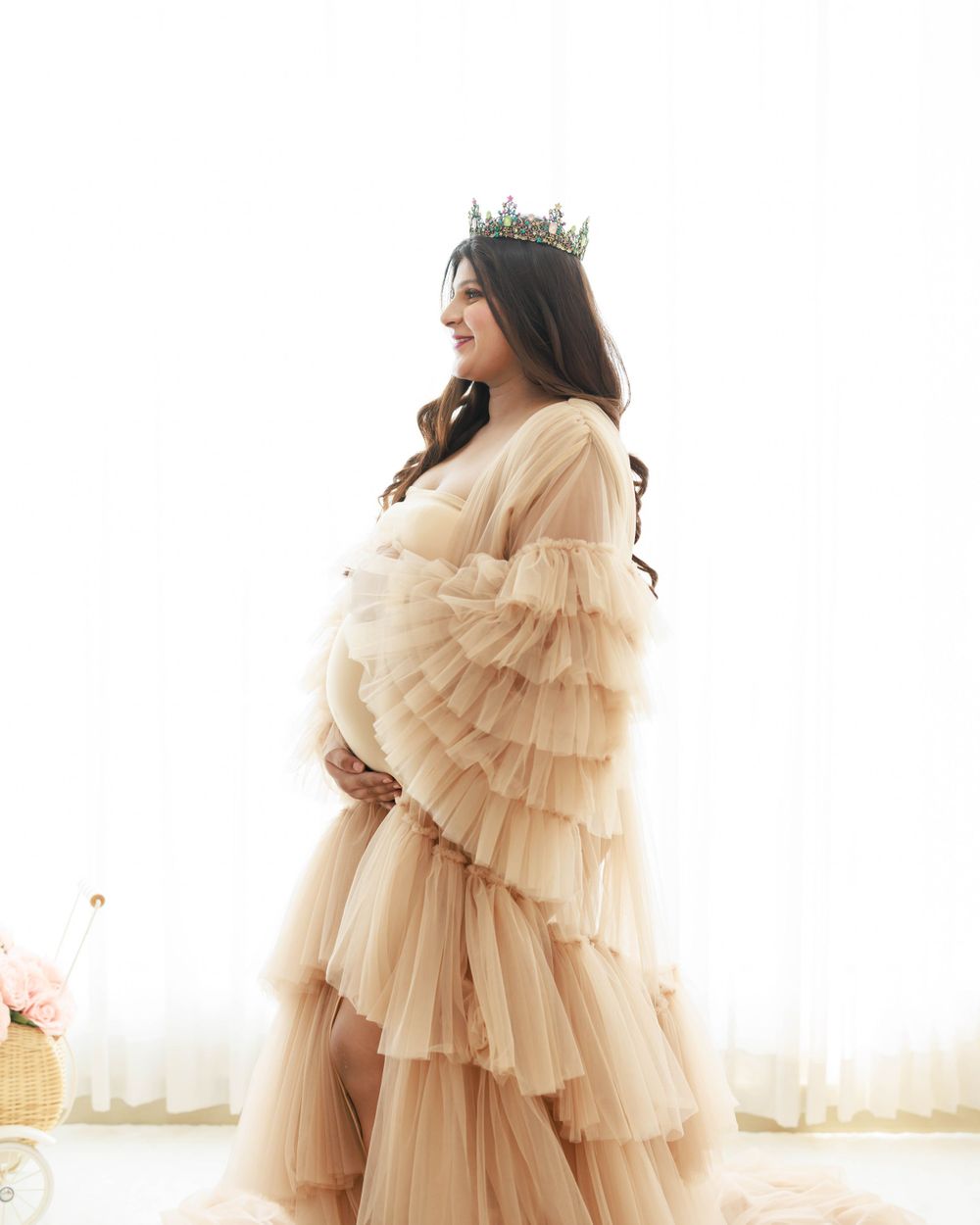 A side view maternity portrait in a ruffled beige dress with a crown accessory.