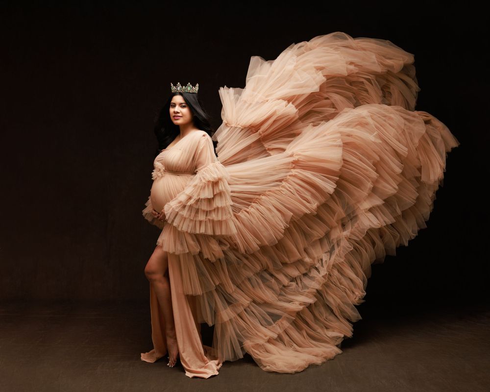 Artistic maternity photo with flowing blush pink ruffled gown against dark background.