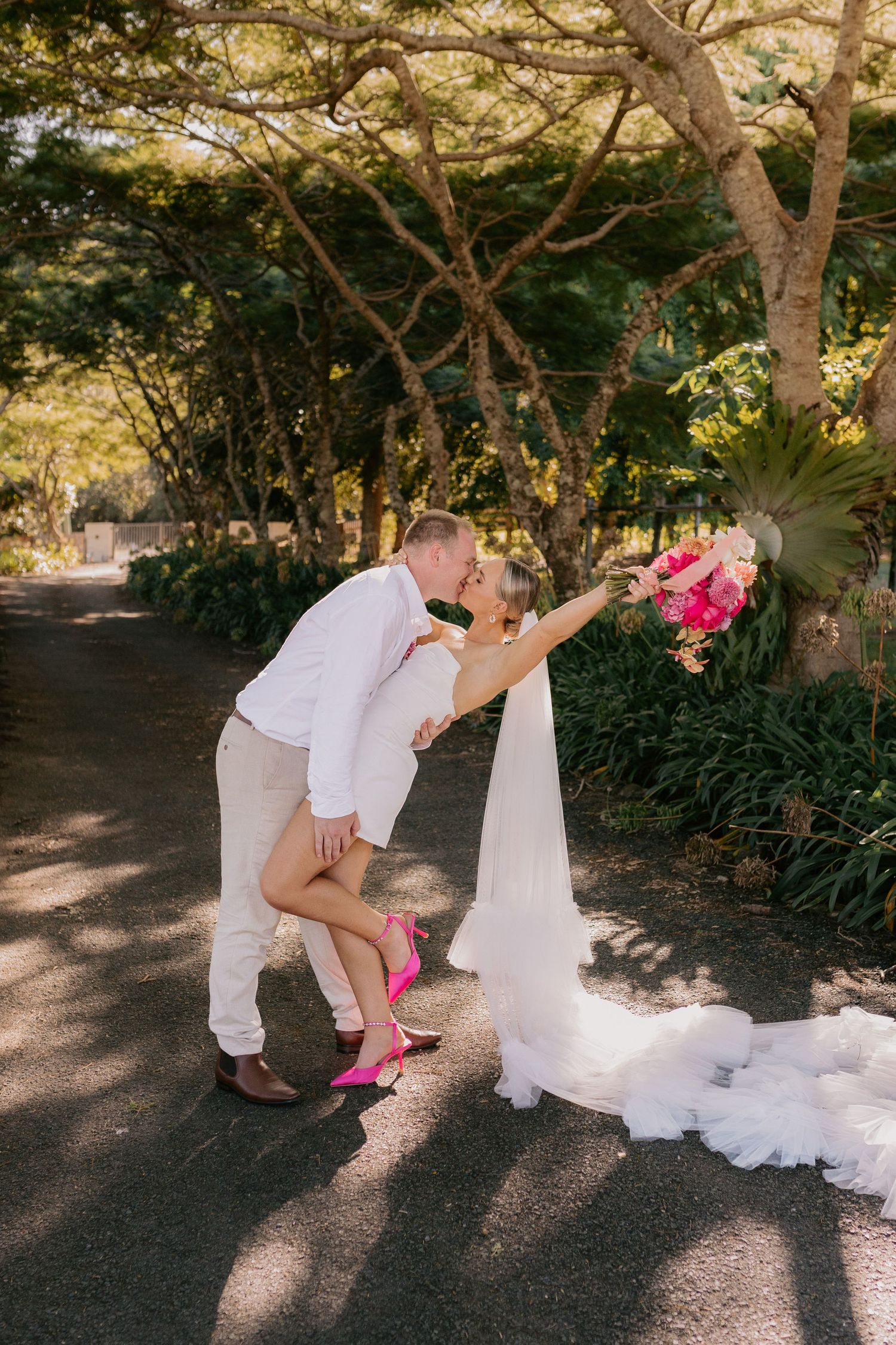 A romantic wedding photo of a couple sharing a kiss on a tree-lined path with flowing white dress and pink bouquet.